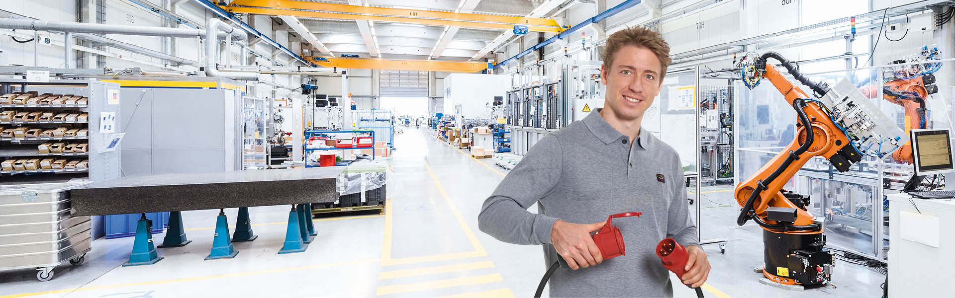 Ein Mann mit grauem Oberteil hält einen roten Stecker und eine rote Kupplung in der Hand. Im Hintergrund ist eine Produktionshalle zu sehen