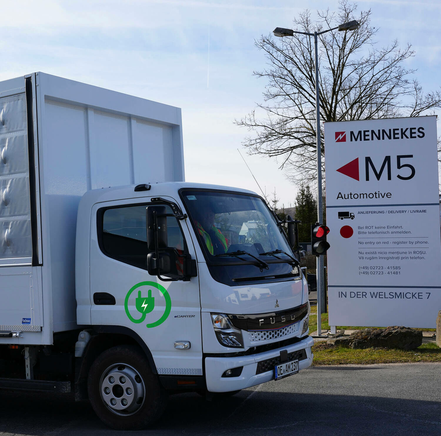 Logistik und Mobilität Weißer Elektro-Lkw mit grünem Steckersymbol passiert MENNEKES-Schild „M5 Automotive“.
